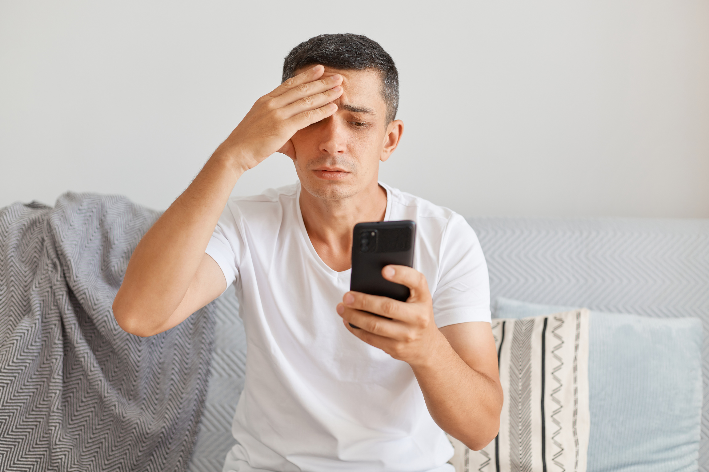 Indoor Shot of Sad Upset Man Wearing Casual Style White T Shirt Sitting on Sofa, Using Mobile Phone with Sorrow Expression, Keeping Hand on Head, Suffering Headache.